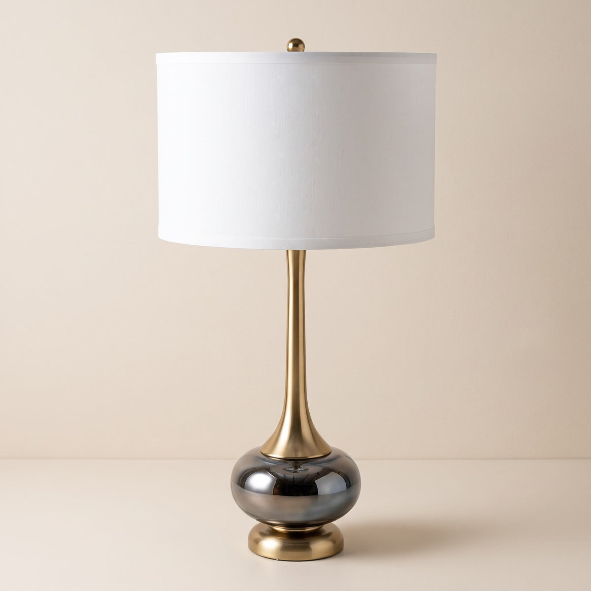 Gray glass and gold table lamp with white drum shade on an ivory backdrop