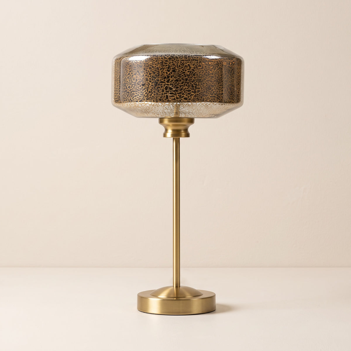 Gold table lamp with mercury glass drum shade and slim brass finish stem on an ivory backdrop