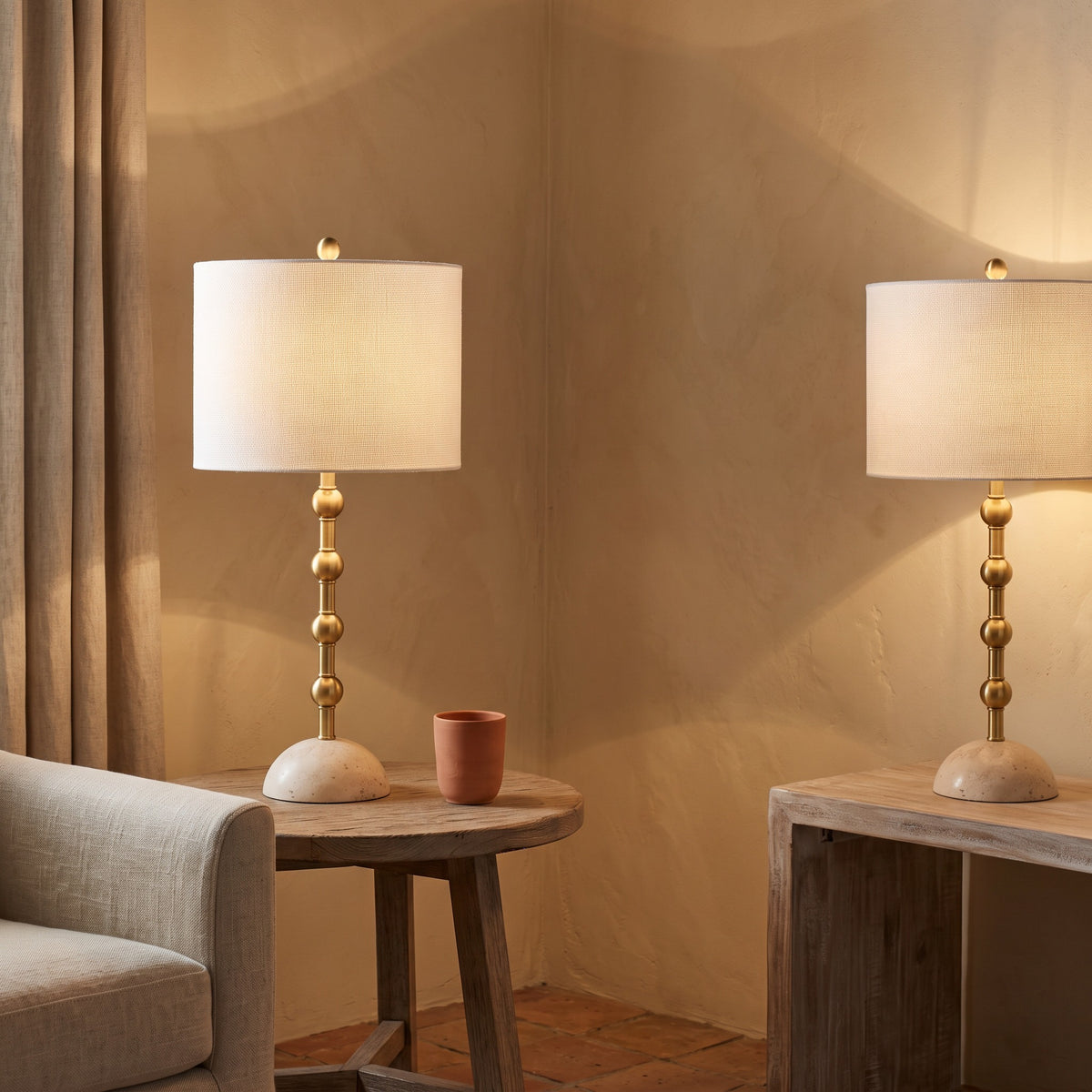 Pair of gold table lamps with travertine bases and off-white shades lit on a side table and console