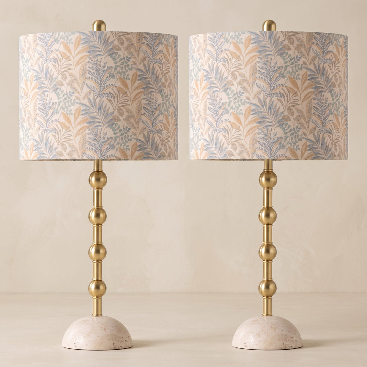 Pair of brass table lamps with travertine bases and leaf print drum shades on an ivory plaster backdrop