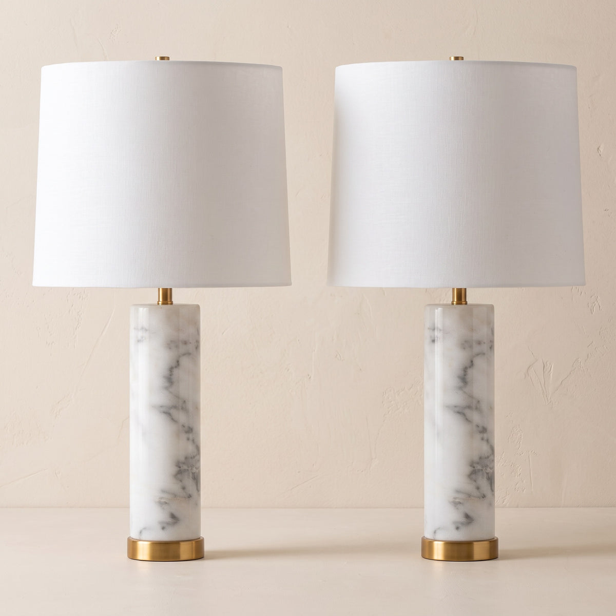 Two white marble table lamps with white linen shades and brass finish bases against an ivory plaster wall