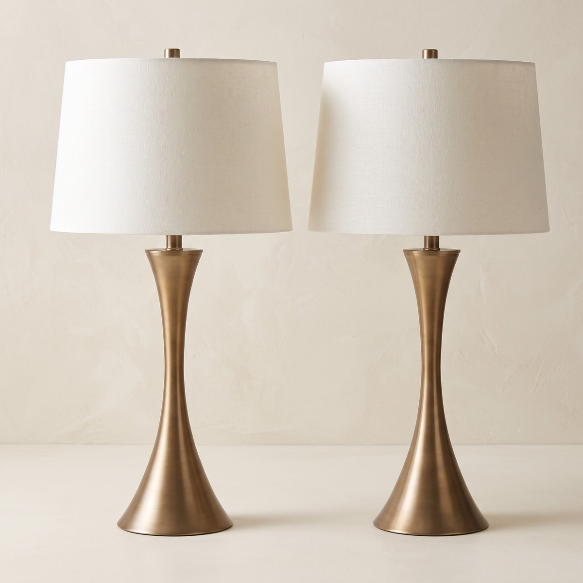Pair of golden bronze hourglass table lamps with off-white linen drum shades on an ivory plaster backdrop
