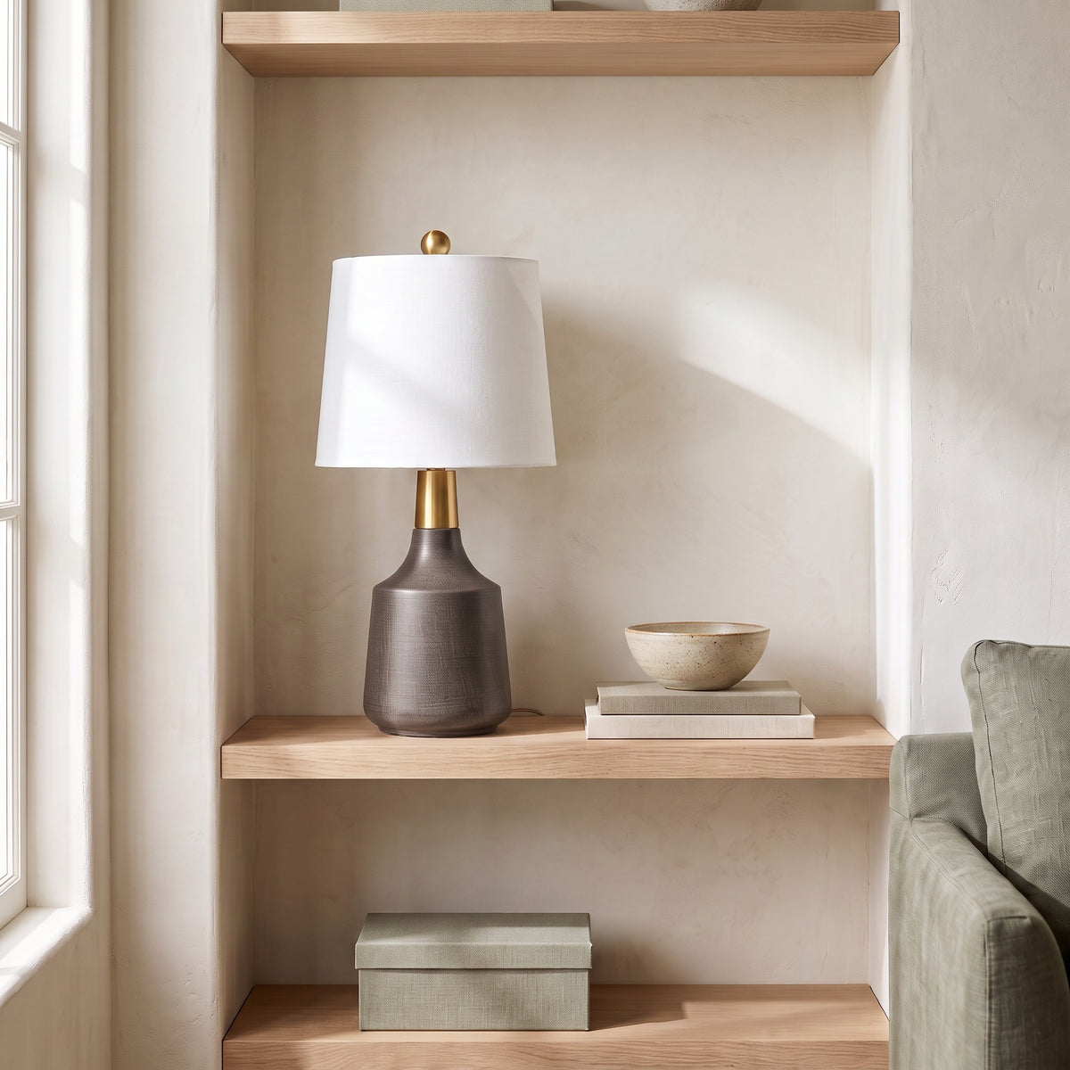 Gray ceramic table lamp with white linen shade and brass neck on an oak shelf beside a ceramic bowl