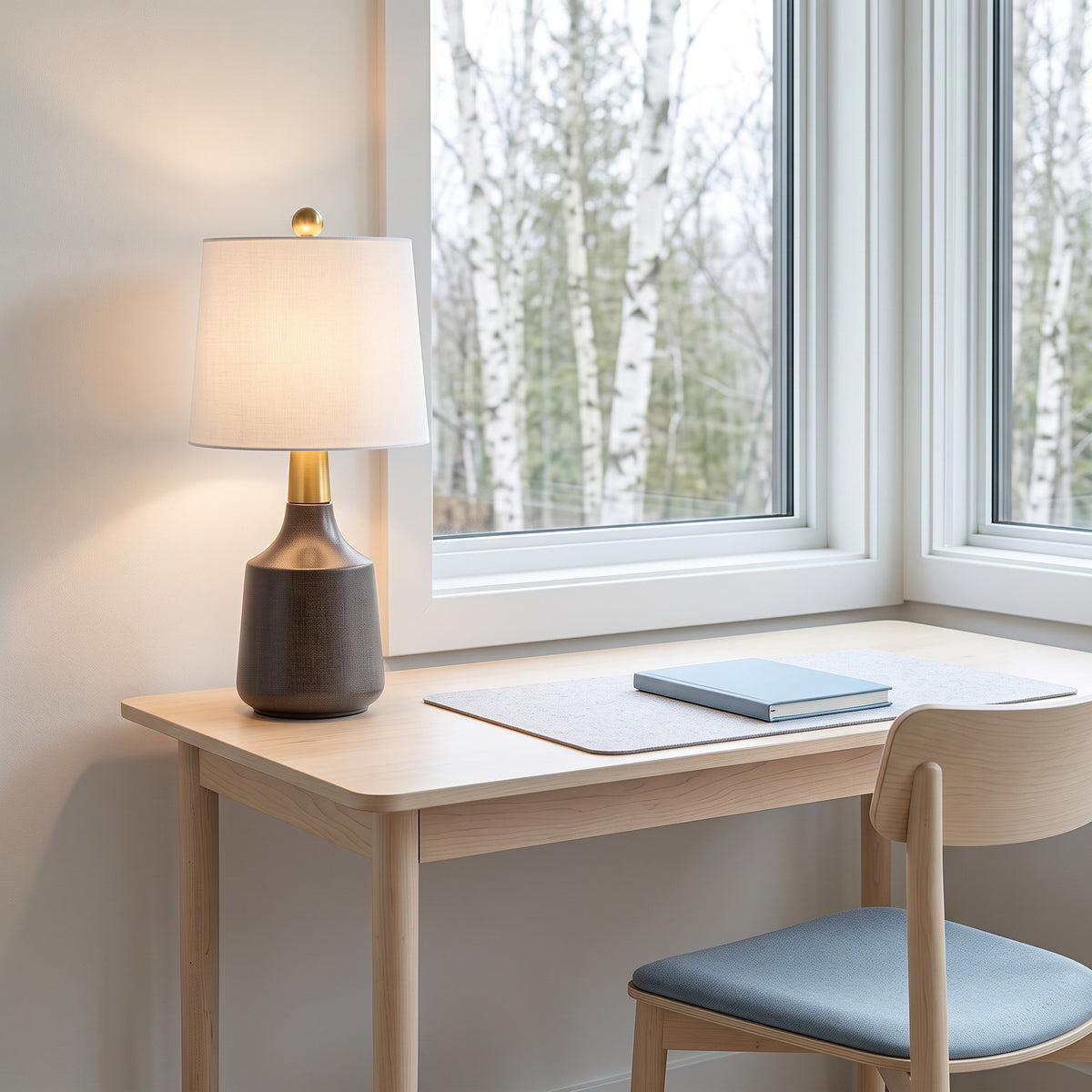 Gray ceramic table lamp with brass neck and lit white linen shade on a light wood desk by a window