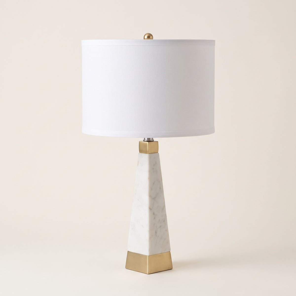 White marble table lamp with brass trim and white fabric drum shade on an ivory backdrop