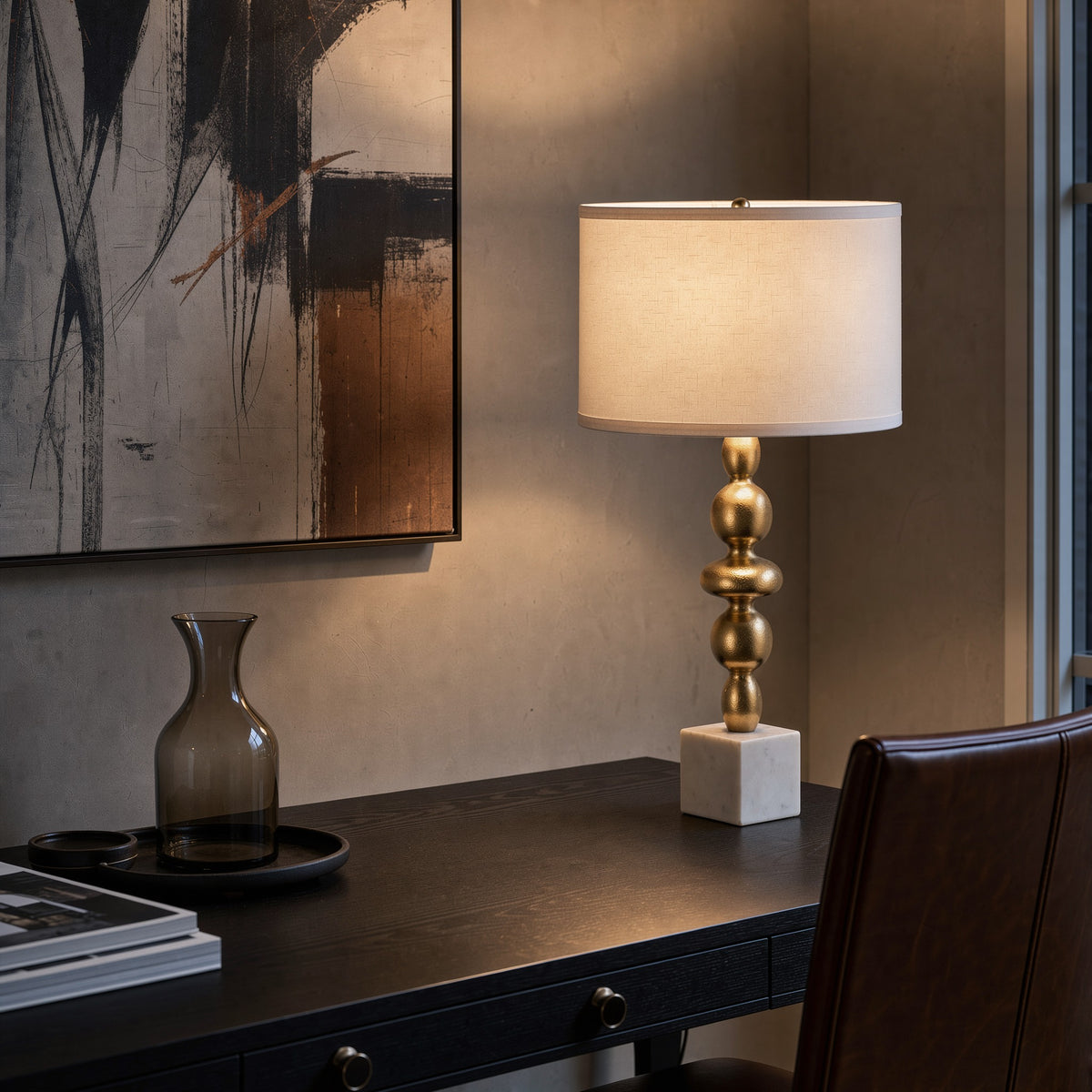 Gold stacked orb table lamp with white marble base and white drum shade lit on a dark wood desk