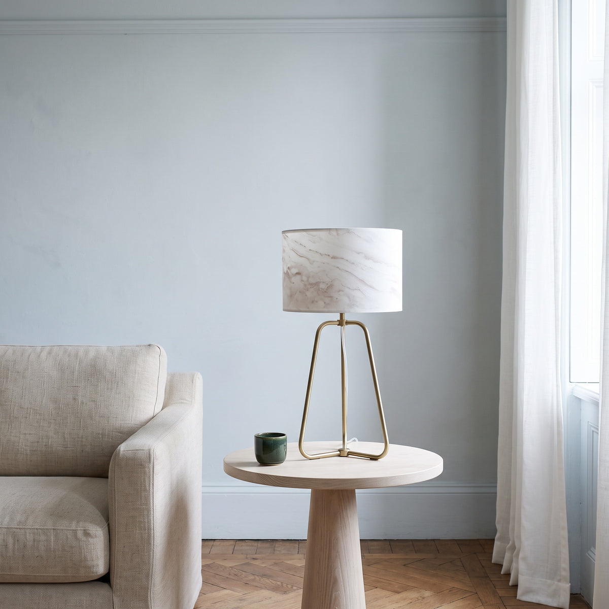 White marble-look drum shade table lamp with brass tripod base on a pale wood side table beside a linen armchair