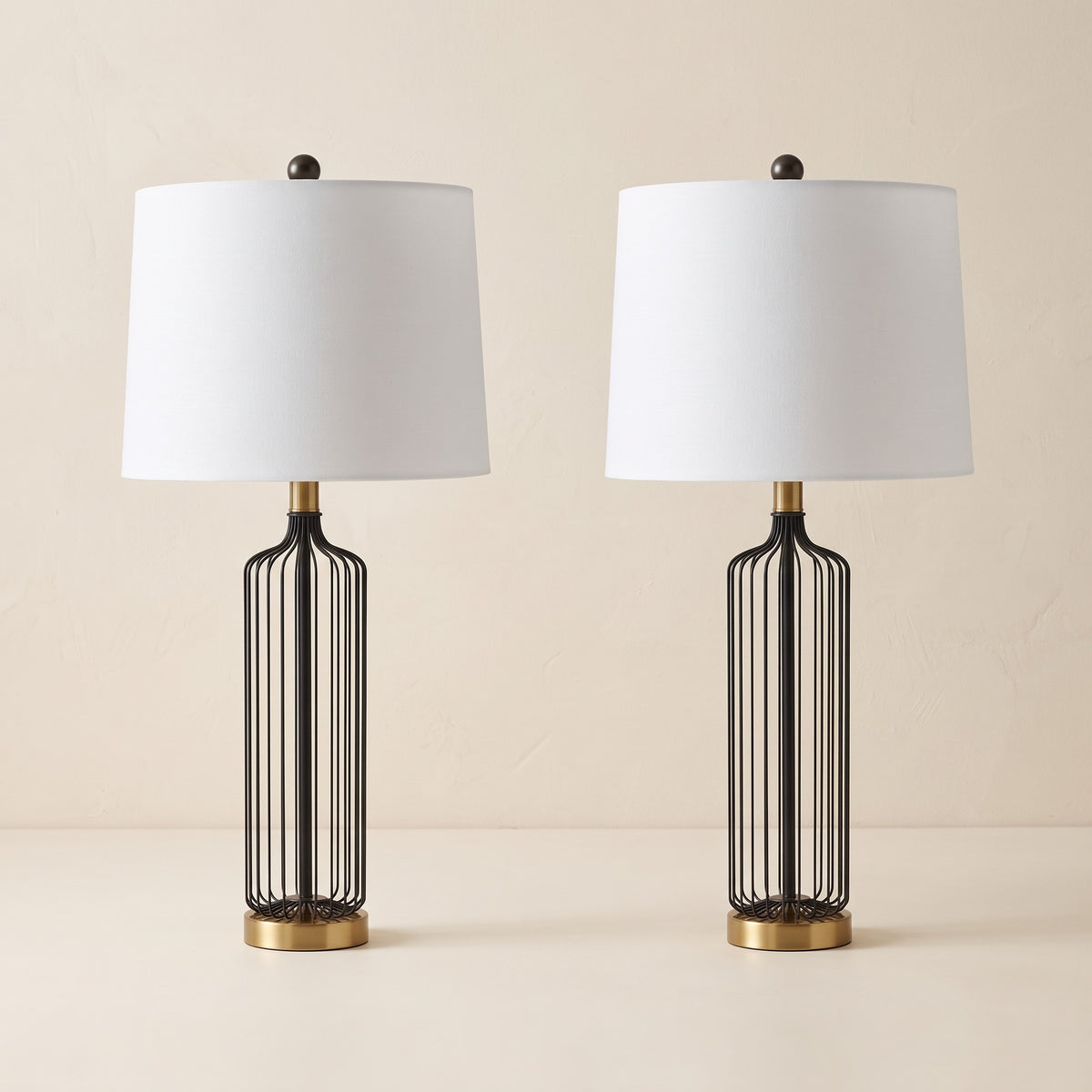 Pair of black wire cage table lamps with brass finish bases and white drum shades on an ivory backdrop