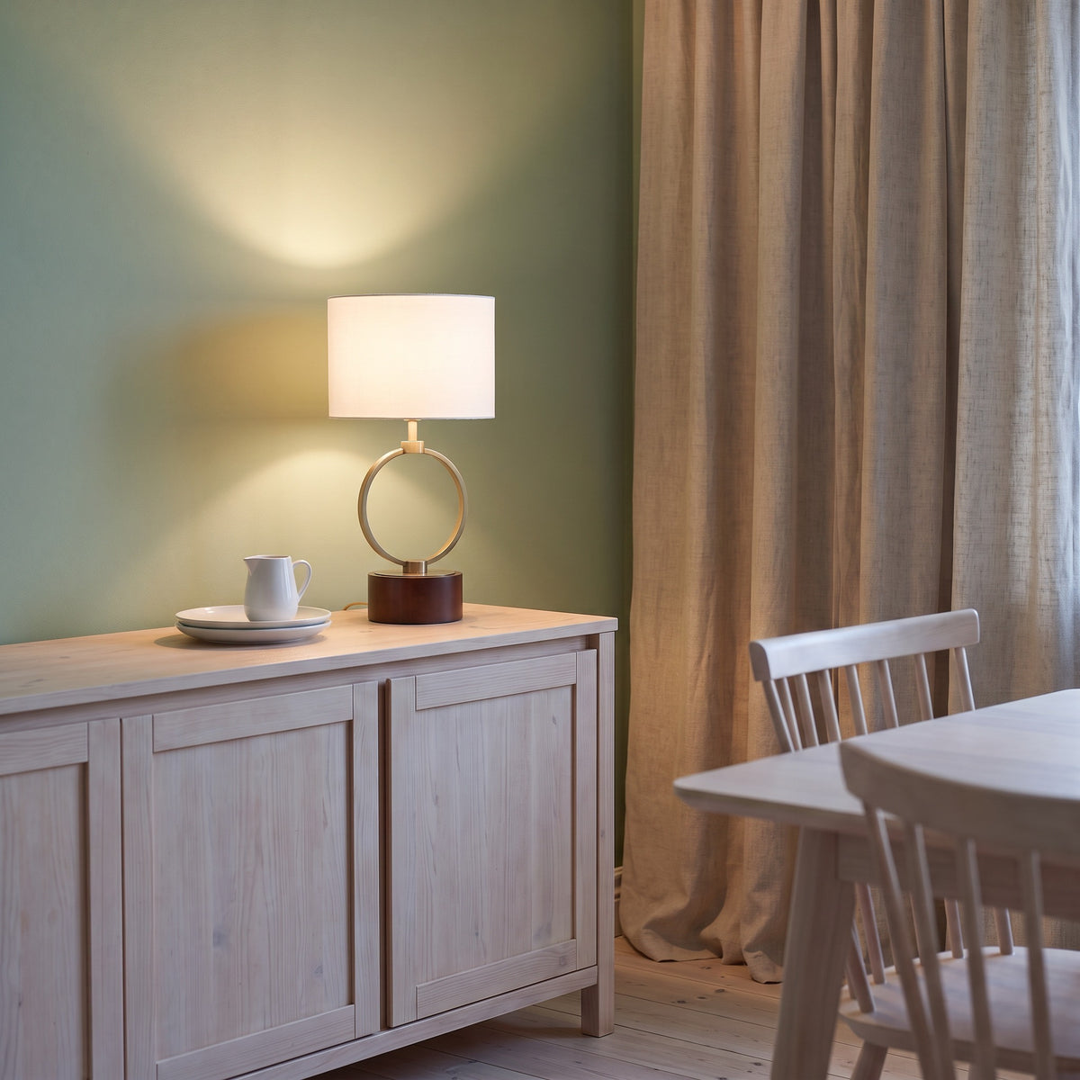 Brass ring table lamp with white linen drum shade and walnut finish wood base, lit on a light wood sideboard