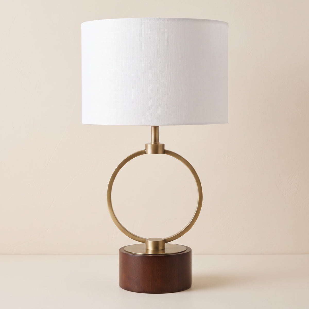 Brass ring table lamp with white linen drum shade and round walnut finish wood base on an ivory backdrop