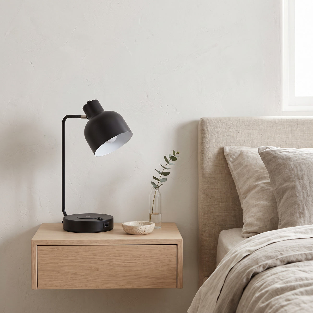 Black metal task table lamp with white inner shade and charging base on an oak floating nightstand by a linen bed