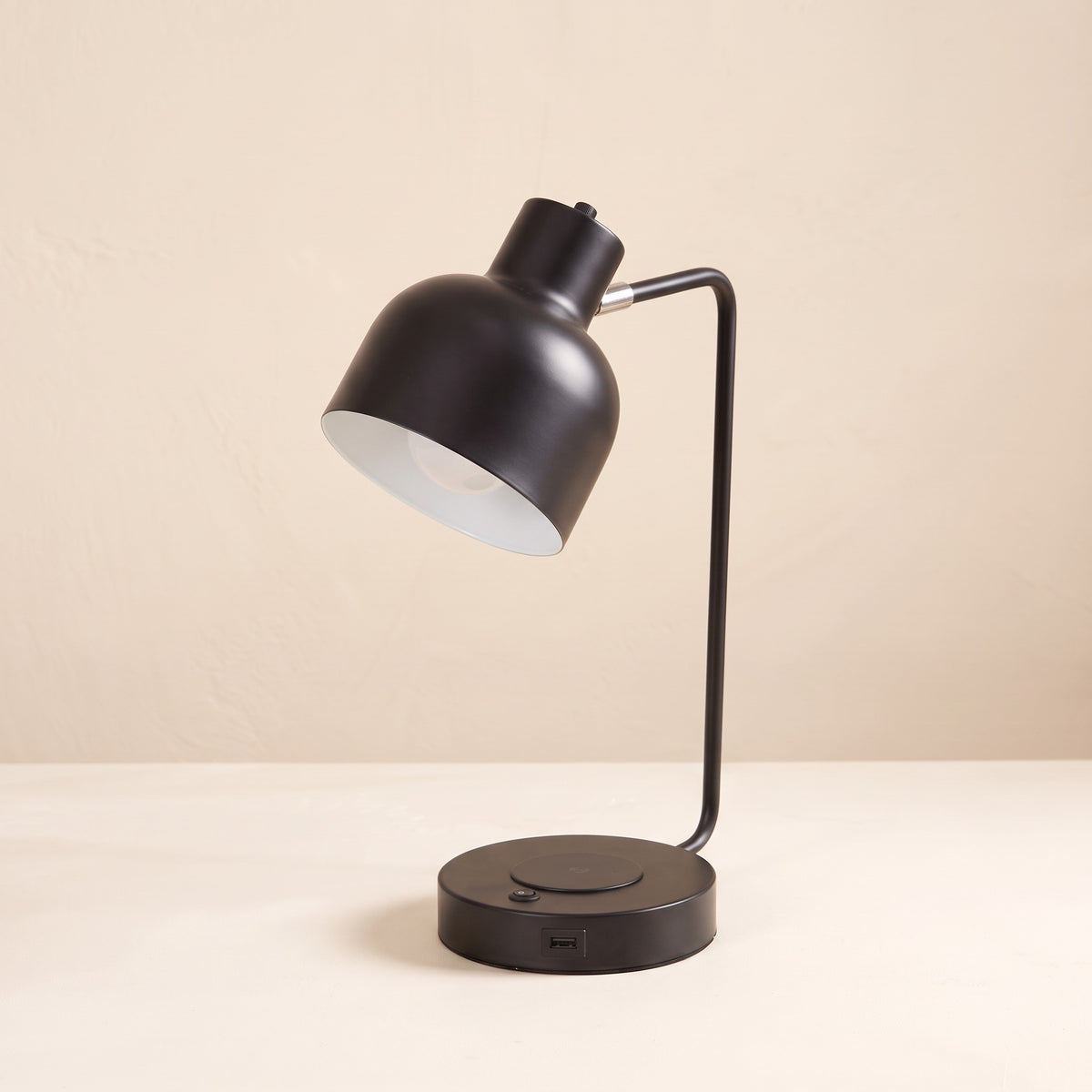 Black metal task table lamp with white lined dome shade and charging pad base on an ivory backdrop
