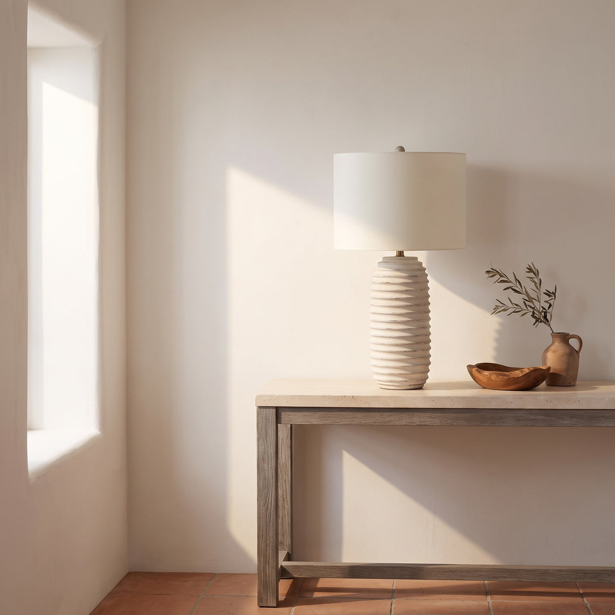 Cream ridged table lamp with ivory linen-look drum shade on a stone-top console beside a sunlit plaster window