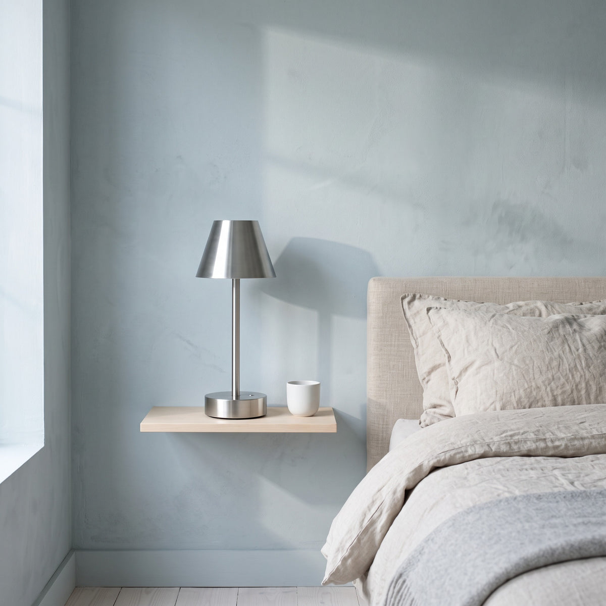 Brushed nickel cordless table lamp with metal cone shade on a floating wood shelf beside a linen bed