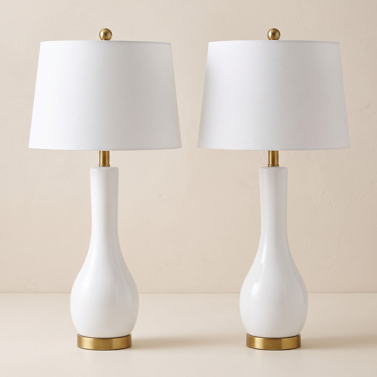 Pair of white ceramic table lamps with white drum shades and brass feet on an ivory backdrop