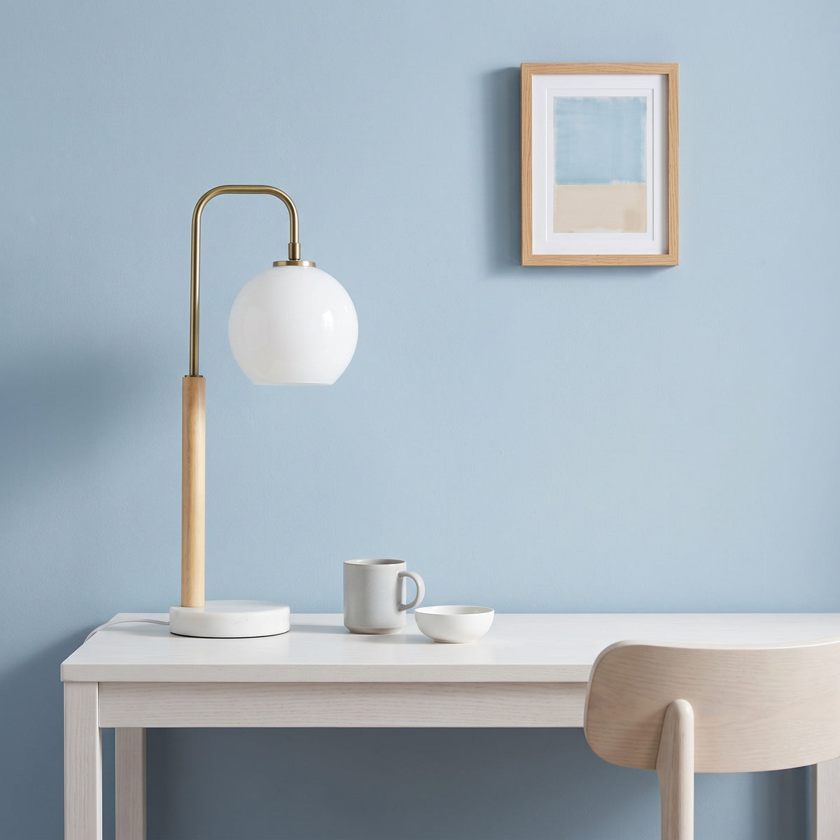White globe table lamp with brass finish arm, light wood stem and marble base on a white desk by a blue wall