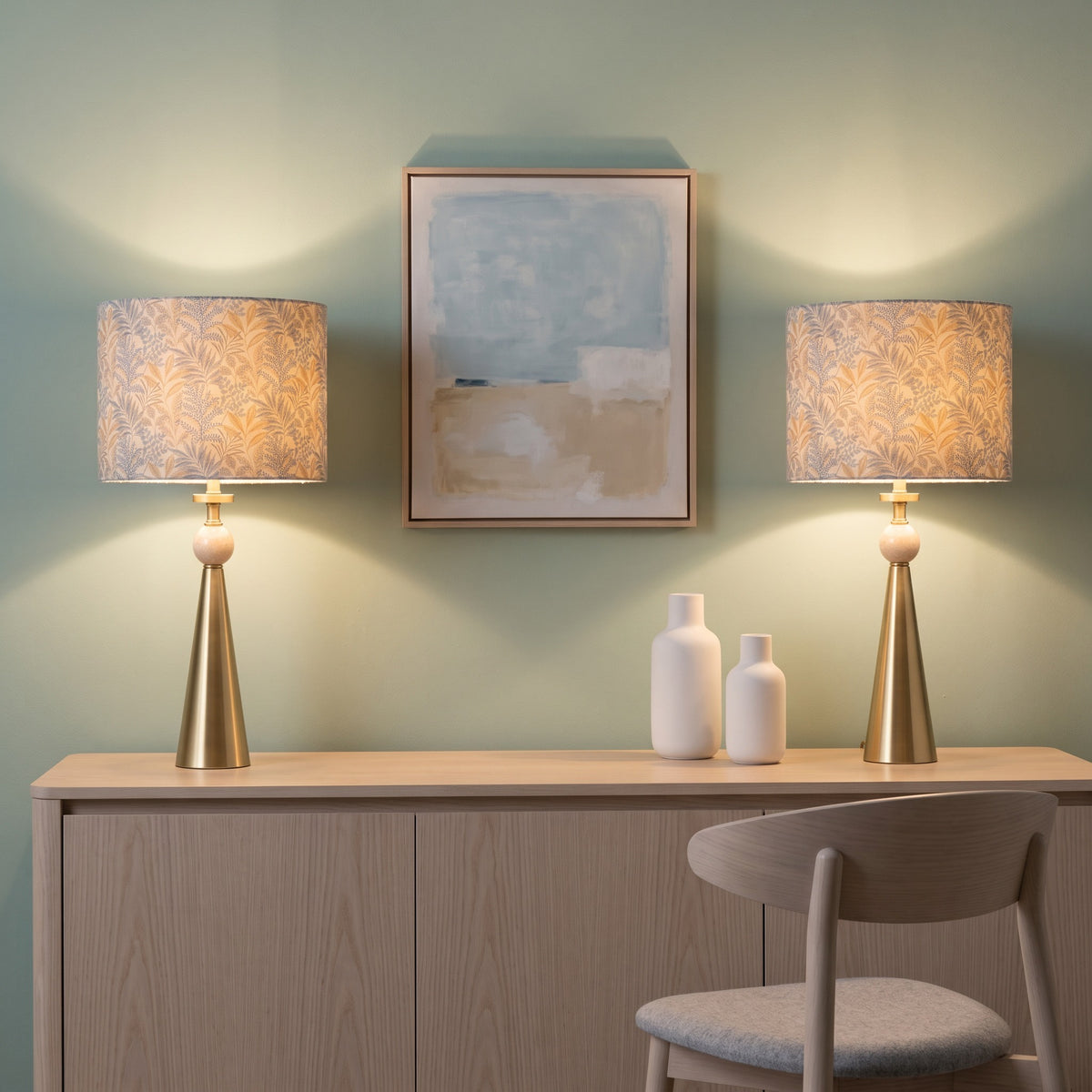 Pair of brass and travertine table lamps with leaf print drum shades, lit on a light wood console