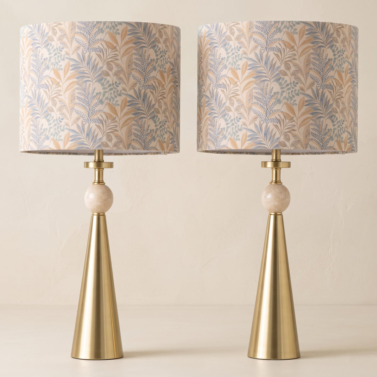 Set of two gold cone table lamps with travertine ball and leaf-print drum shades on an ivory backdrop