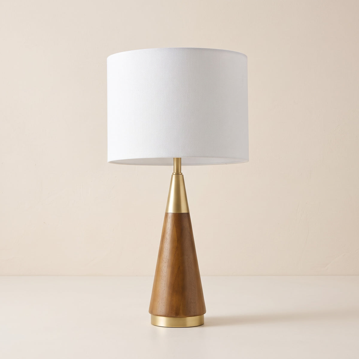 Table lamp with brown wood cone base, brass accents and white linen drum shade on an ivory backdrop