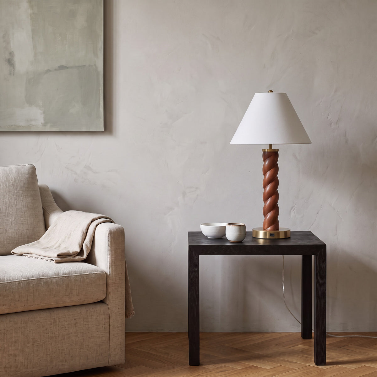 Table lamp with twisted wood-look column, brass finish base and white shade on a dark side table by a linen armchair