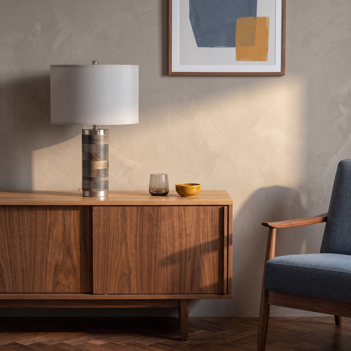 Wood table lamp with striped grey and tan base and gray drum shade on a walnut sideboard beside a blue armchair