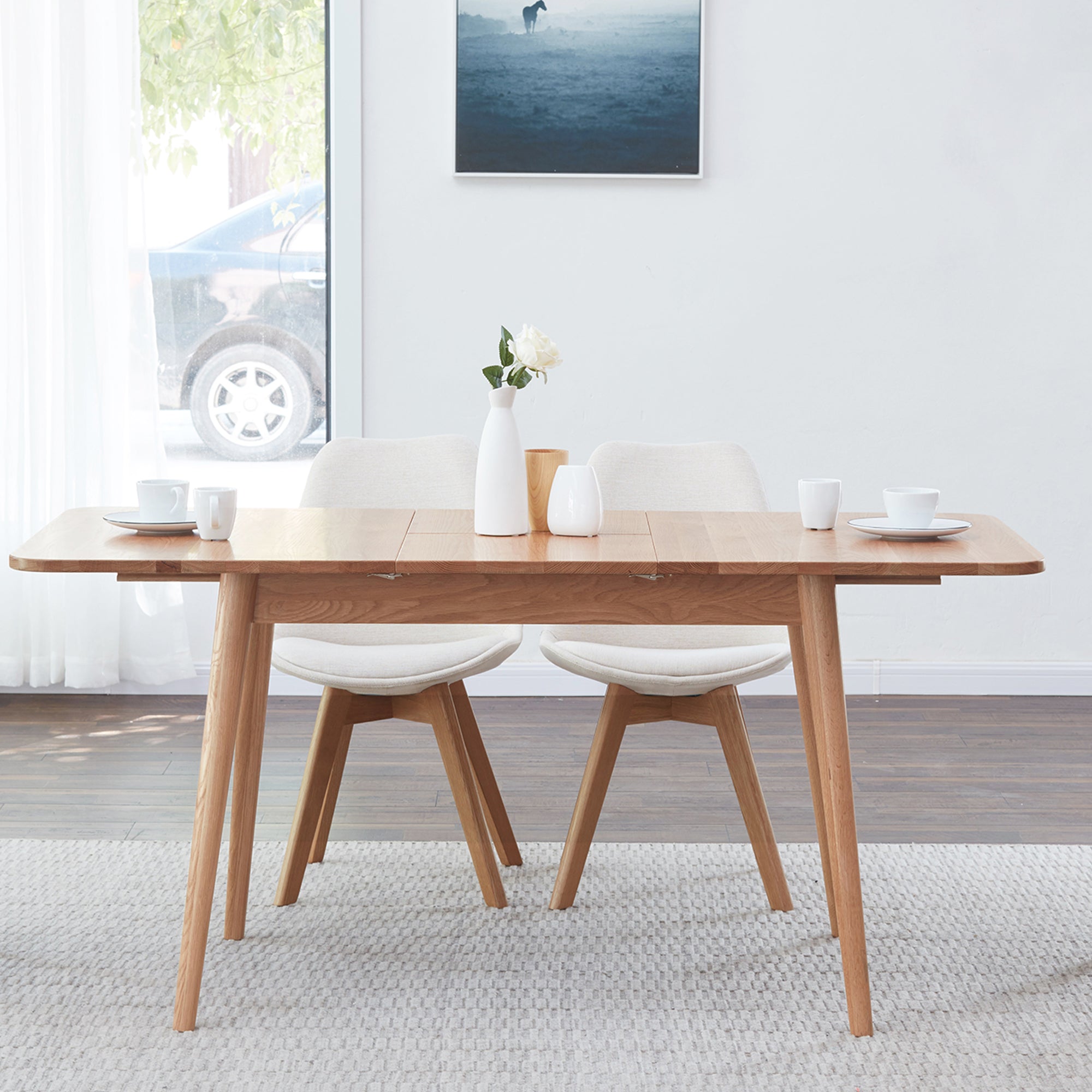 Natural oak dining table with two cream chairs by a window, cups and a white vase on top