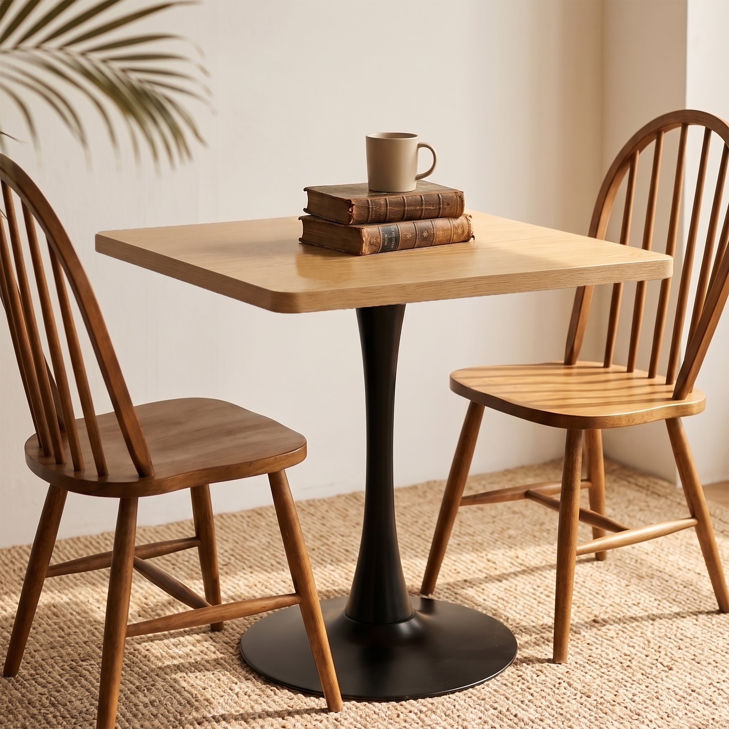 Natural square tulip dining table flanked by two wood Windsor chairs with stacked books and a mug