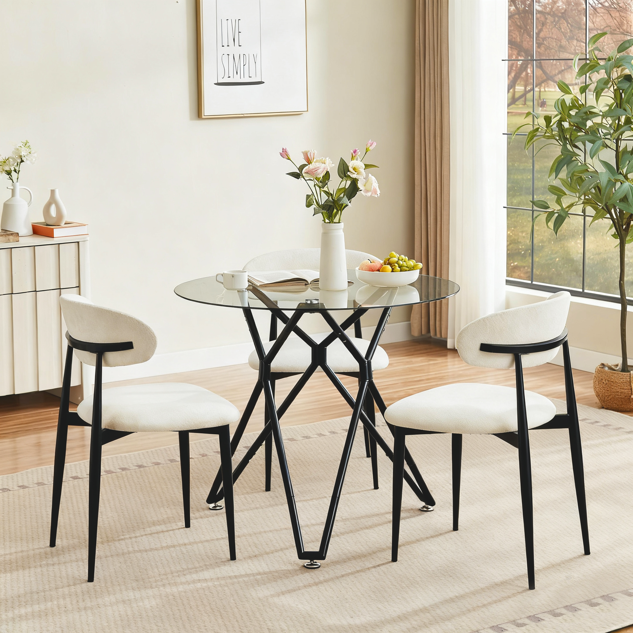 Round glass dining table with three cream boucle chairs below a Live Simply print