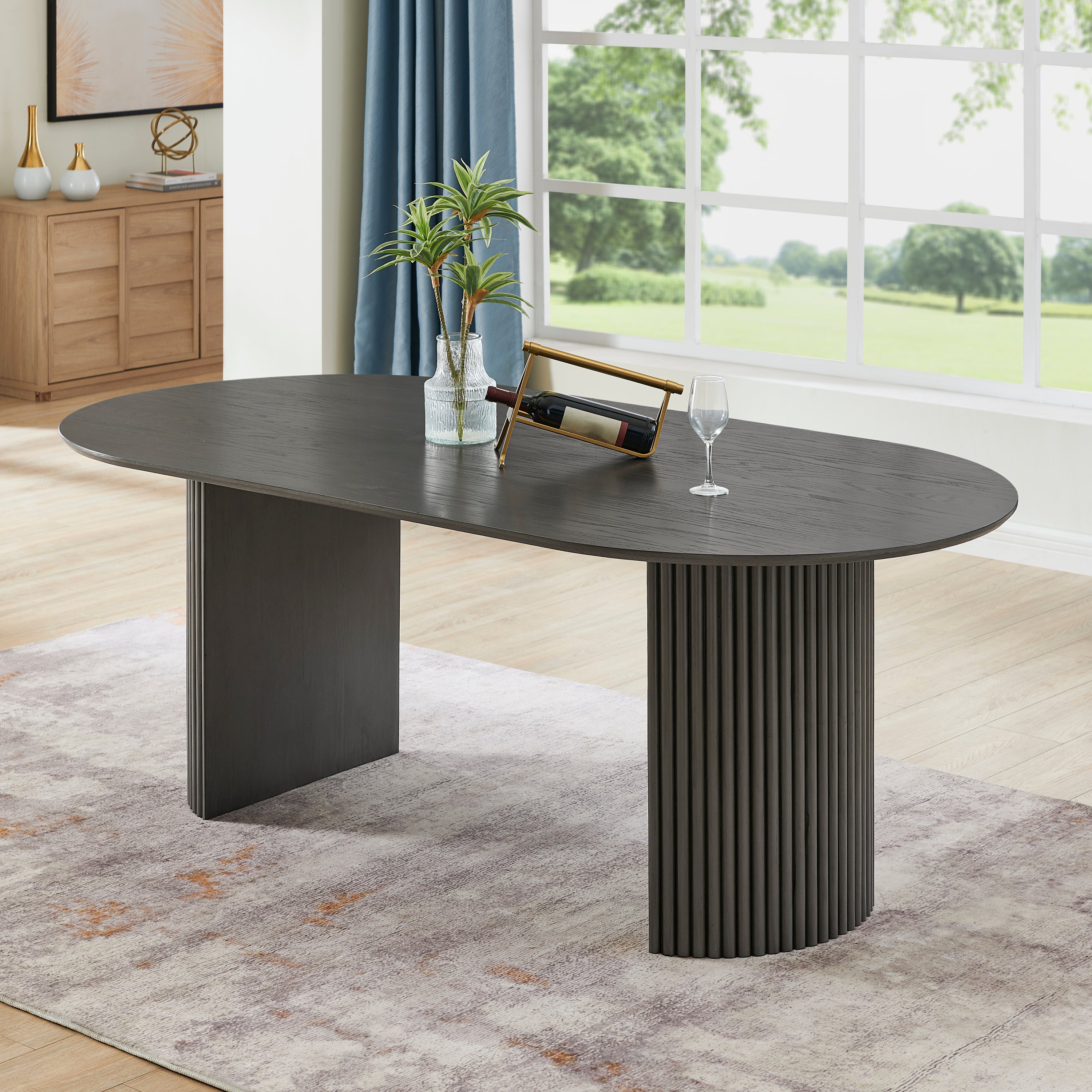 Gray oak oval dining table with fluted pedestal base in a bright living room, styled with wine and a plant